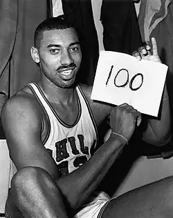 Wilt Chamberlain, an African American man, is shown sitting down in his Philadelphia Warriors jersey while holding up a piece of paper with the number 100 written on it. The photograph was taken directly after the game and is in black and white.