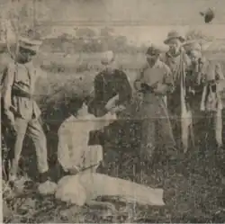 A grainy image of a man, accompanied by five others, leaning over the body of a woman