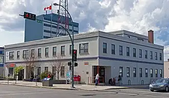 A two-story light gray building seen from across an intersection over which a traffic signal is displaying a red light. A Canadian flag is flying from the flat roof. There are taller buildings behind it.