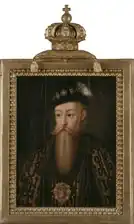 A man with a long blond beard in an ornate gold and black tunic wearing an equally ornate hat with a large white feather in it and a large bejeweled religious icon around his neck