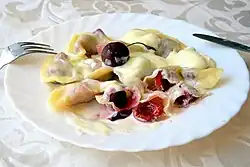 Duży talerz z porcją pierogów z wiśniami. Niektóre pierogi przekrojono na pół, odsłaniając nadzienie. Danie polane jest sosem, prawdopodobnie waniliowym. Całość zwieńczona jest pośrodku jedną wiśnią z ogonkiem. Wiśnie o ciemnoczerwonej barwie.