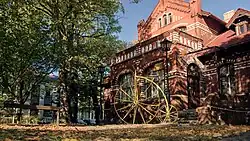 Fotografia barwna, willa z czerwonej cegły, a przed nią żółta rzeźba zapewne z metalu w kształcie bicykla oraz z lewej drzewa liściaste