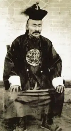 Forografia czarno-biała, przedstawia siedzącego mongolskiego dygnitarza w średnim wieku w regionalnym ubiorze i mongolskiej czapce na głowie, na ubraniu na piersiach ma tradycyjny okrągły mongolski symbol