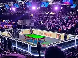 Zdjęcie z turnieju. Stół snookerowy otoczony trybunami pełnymi widzów.