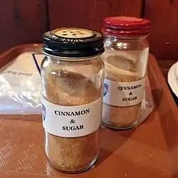 Dwa opakowania cukru z cynamonem. Widoczne etykiety z angielską nazwą „cinnamon & sugar”. Szklane, wąskie słoiczki z metalowymi zakrętkami, w których widoczne są niewielkie otwory, przez które dozuje się przyprawę. Zakrętki w dwóch różnych kolorach: czarnym i czerwonym
