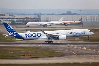 Zdjęcie samolotu na płycie. Duży pasażerski z uniesionymi końcówkami skrzydeł. Duży napis 1000 na burcie.