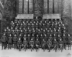 Zdjęcie dużej grupy żołnierzy w mundurach wojskowych na tle dużego oszklonego budynku. Mężczyźni w pierwszym rzędzie siedzą, a w pozostałych sześciu rzędach stoją na platformach.