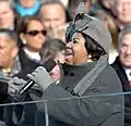 A woman her mouth open, holding a microphone. She is wearing a gray hat with a large bow, gray gloves, and a gray jacket. In the background is a crowd of people.