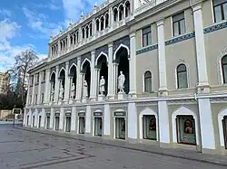 Zdjęcie wykonane w słoneczny dzień przedstawia fasadę budynku muzeum. Fasada sfotografowana jest z bliska, ukośnie. Utrzymana jest w jasnej kolorystyce, podzielona rytmicznie ułożonymi pilastrami. W centralnej części znajduje się sześć nisz zamkniętych łukami. W każdej z nich jest umieszczony posąg.