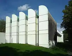 Elewacja budynku muzeum i archiwum składająca się z pięciu jednakowych łukowato zakończonych elementów pośród zieleni