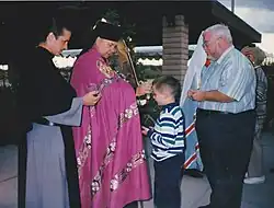 Duchowny ubrany w szaty w stylu bizantyjskim: czarny kłobuk, fioletowy płaszcz czyni znak krzyża na czole chłopca. Biskupowi towarzyszy ksiądz w szarym habicie z czarnym szkaplerzem, a za chłopcem stoi starszy mężczyzna.