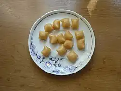 Porcelanowy, zdobiony, spodek, na którym leży kilkanaście beżowych kostek podobnego rozmiaru. Wszystkie mają kształt zbliżony do grubej poduszki.