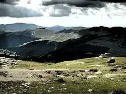 Zdjęcie krajobrazowe. U dołu – fragment szczytu górskiego, z którego wykonano zdjęcie, w większości porośnięty trawą. W centralnej części sąsiedni masyw – góry do połowy wysokości zalesione, wyżej trawiaste. Za nim dalsze szczyty. W tle zachmurzone niebo.