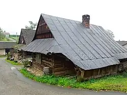 Zdjęcie przedstawia tradycyjną, drewnianą chałupę. Budynek ustawiony jest szczytem do drogi, nakryty wysokim dachem półszczytowym. Boczna połać dachu jest przedłużona na przylegające do budynku dobudówki, od strony drogi tworzy rodzaj podcienia. Chałupa jest przy asfaltowej drodze, na terenie pokrytym trawą. Za nią widoczny jest fragment drugiej, podobnej chałupy, a w tle za nimi fragment stodoły.