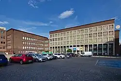 Fotografia barwna, prosta modernistyczna fasada z żelbetu i cegły w układzie kratownicy z prawej, a z lewej prosta ceglana fasada, oba budynki połączone ceglaną przewiązką