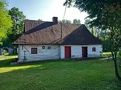 Jednokondygnacyjny, biały budynek, z dachem półszczytowym. Lewa część budynku jest drewniana, z jednym oknem. Prawa część budynku ceglana, otynkowana. W tej części jest dwoje drzwi, jedne zakratowane. Obok drzwi jest okienko. Na szczycie dachu, w środku długości domu, jest ceglany komin. Dom stoi na niskim, fundamencie. Wokół domu rozciąga się trawnik. W tle drzewa, a po lewej stronie fragment bramy.
