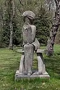Zdjęcie rzeźby parkowej wykonanej ze sztucznego kamienia. Przedstawia siedzącą na ławce młodą kobietę. Kobieta jest szczupła, ubrana w wąską sukienkę do kolan. Ręce trzyma oparte na złączonych kolanach, palcami podpiera brodę. Włosy proste, zakrywają uszy. Uwagę przyciąga głowa - w miejscu twarzy jest prosta płaszczyzna z pionową szczeliną pośrodku. Wokół rzeźby trawnik, z tyłu drzewa.