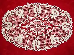 Owalna, ażurowa szydełkowa serweta na czerwony tle. Widoczne są motywy kwiatowe połączone z siatkowym tłem, również wykonanym na szydełku.