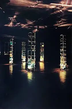 Kolorowa fotografia. Ciemne, słabo oświetlone pomieszczenie z multimedialną instalacją artystyczną. Głównymi elementami instalacji widocznymi na fotografii jest pięć kolumn – pionowych konstrukcji wznoszących się od podłogi niemal do sufitu – są one wąskie, czworościenne, ściany mają zbudowane z mniejszych części-obrazów. Kolumny są nieregularnie podświetlone od środka – część ich elementów jest oświetlona kolorem żółtym, część zielonym, a część z nich jest pogrążona w mroku. Podłoga pomieszczenia również znajduje się w ciemności – jedynie niewielkie fragmenty w okolicach kolumn są oświetlone światłem z nich pochodzącym. Na ciemnym suficie widać nieregularne jaśniejsze kształty, rzucane przez oświetlenie instalacji. Tylne ściany pomieszczenia giną w mroku.