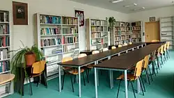 Na środku owalny, drewniany stół z kilkunastoma krzesłami. W tle regały z książkami i godło Polski. Zielona wykładzina na podłodze.