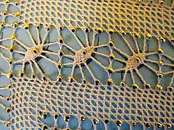 Zdjęcie – zbliżenie na fragment koronki. Wzór to tzw. pajączki – elementy o kształcie łezki z rozwidlającymi się nićmi, rozpięte pomiędzy przypominającymi siatkę pasami. W koronkę wkłute są szpilki, aby umocować i ustabilizować ją w trakcie wyplatania.