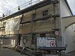 Zdjęcie przedstawia budynek w czasie remontu sfotografowany z ukosa. Przy ścianie domu stoją rusztowania, na ulicy, przy rusztowaniu, są położone bezładnie belki i drabina. Cześć rusztowania zasłonięta jest tkaniną, być może jest to reklama. Na rusztowaniu znajduje się kilka osób.