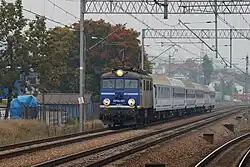 Na zdjęciu widać zbliżający się do widza pociąg z wagonami przedziałowymi na przedzie którego jedzie niebieska lokomotywa z włączonymi lampami. Obok kolejne tory, nad nią przewody elektryczne.