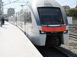 Zdjęcie. Czoło nowoczesnego elektrycznego zespołu trakcyjnego, stojącego przy krawędzi peronu. Widoczny bok pociągu, znajdujący się od strony peronu. Zespół trakcyjny składa się z trzech dwupiętrowych członów. Pomalowany jest na biało, z ciemnoszarą, otoczoną złotym paskiem dekoracją w postaci szerokiego, ciągnącego się przez całą długość boku pociągu pasa, w połowie wysokości pociągu oraz z szarym ozdobnym paskiem przy dachu, rozszerzającym się na czoło pociągu aż do wysokości dolnej krawędzi przedniej szyby. Pod szybą na czole pomarańczowa dekoracja. Widoczny napis STADLER na boku kabiny maszynisty.