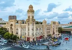 Zdjęcie. W centralnej części widoczny w rzucie izometrycznym budynek dworca. Przed nim plac z kilkudziesięcioma poruszającymi się ludźmi, kioskiem, zejściem na stację metra. Po lewej stronie zdjęcia przed budynkiem kilkupasmowa ulica z samochodami zatrzymanymi przed widocznym u dołu fotografii przejściem dla pieszych, odgrodzona od budynku szpalerem drzew. W tle niebo z chmurami. Budynek wykonany z kamienia o barwie jasnego piasku, posiadający trzy wysokie piętra z licznymi oknami. Bryła prostopadłościenna z licznymi zdobieniami. W połowie każdej ściany wywyższony ponad bryłę budynku prostokątny portal z prowadzącymi na wysokość pierwszego piętra szerokimi schodami. W rogu budynku, widocznym w środkowej części fotografii, ozdobna wieża na planie kwadratu, o obłym zakończeniu, około dwukrotnie wyższa od bryły budynku. W połowie odległości pomiędzy narożnikami budynku a portalami umieszczone ozdobne wieżyczki na planie kwadratu, wysunięte nieco poza bryłę budynku, o wysokości portalu, o obłym zakończeniu.