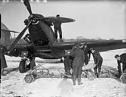 Czarno biała fotografia samolotu stojącego na śniegu śmigłem do oglądającego. Przed prawym skrzydłem samolotu stoi grupa żołnierzy. Dwóch z nich trzyma bombę o długości zbliżonej do wzrostu żołnierzy. Dwaj żołnierze obserwują lub nadzorują niosących bombę a trzeci manipuluje przy podłużnym wózku z którego podniesiono bombę. Na prawym skrzydle samolotu stoi mężczyzna zasłonięty częściowo przez śmigło samolotu. W tle widać częściowo zasłonięte przez samolot drzewa na horyzoncie.