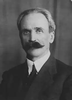 Starszy mężczyzna w garniturze, ma siwe włosy i duże czarne wąsy. Ma pogodny wzrok, lekko się uśmiecha.