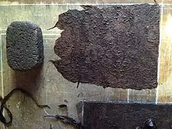 Zbliżenie na narzędzia do produkcji papieru amate: na płaskiej desce oznaczonej płytkimi żłobkami wyznaczającymi krawędź arkusza około jednej czwartej powierzchni zajmuje wilgotna miazga kory. Widoczny również sześcienny kawałek wulkanicznej skały używany do rozdrobnienia.