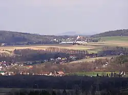 Zdjęcie przedstawia górski krajobraz sfotografowany ze stoku. Kadr wypełniają niskie góry o łagodnych zboczach pokryte lasami, łąkami i polami. W dolinach między pagórkami znajdują się zabudowania wsi - niskie domy z dwuspadowymi dachami. Wśród nich wyróżnia się budynek kościoła z dominującą wieżą.