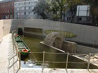 Zdjęcie zrobione w słoneczny dzień przedstawia wylot kanału. Na pierwszym planie jest zbiornik wodny obudowany betonową ścianą, po lewej stronie, przy nabrzeżu przycumowano długą, zieloną łódź. Na środku zbiornika jest wyspa z wolnostojącą kamienną bramą – to dawny wylot kanału. W tle, widoczny otwór z zawieszonym nad nim godłem górniczym i napisem Szczęść Boże. To obecny wylot kanału