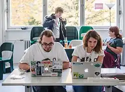 Na pierwszym planie kobieta i mężczyzna siedzą przodem do fotografa, za stołem i oklejonymi rozmaitymi nalepkami laptopami. Na drugim planie kolejne dwie osoby, jedna stoi i śmieje się, druga siedzi obok niej w fotelu. W tle rozświetlone okno.