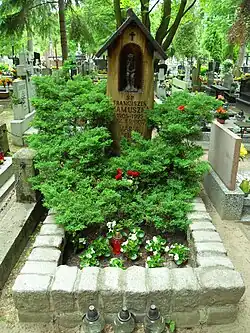 Zdjęcie grobu. Obramowany jest kamiennym murkiem, na części nagrobnej rosną iglaste krzewy oraz fiołki, zaś za nimi znajduje się drewniana kapliczka. Kapliczka to wydrążony pień z rzeźbą Chrystusa Frasobliwego. Na pniu wyrzeźbione są napisy: „ŚP Franciszek Mamuszka, 1905-1995”, (niżej nieczytelny fragment napisu), „Maria Mamuszka” (nieczytelne daty).