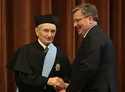 Zdjęcie. Prezydent Polski Bronisław Komorowski ściska dłoń profesora Henryka Samsonowicza. Profesor ma założoną czarną togę akademicką, na głowie nosi biret. W togę wpięty ma order, a przez lewe ramię przerzuconą błękitną wstęgę orderu.