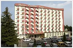 Zdjęcie przedstawia budynek hotelu Cristal Park. Budynek jest wysoki, pomalowany na biało i czerwono. W bryle budowli znajduje się wiele okien i małych balkonów. Po lewej stronie budynku rośnie wysokie iglaste drzewo, a na ziemi widoczny jest parking pełen samochodów.
