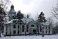 Zdjęcie przedstawia długi, piętrowy budynek nakryty czterospadowym dachem. Po lewej stronie, z dachu wyrasta wieża, nakryta dachem wieżowym z kopułką w zwieńczeniu. W dłuższej elewacji zwraca uwagę trójkątny szczyt, akcentujący główne wejście do budynku. Zdjęcie wykonane jest w zimie. Teren wokół budynku pokryty jest śniegiem. Drzewa przy budynku są bez liści.