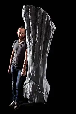 Zdjęcie portretowe. Młody mężczyzna z ciemnymi włosami. Stoi oparty o marmurową rzeźbę, przedstawiającą ramię z zaciśniętą pięścią pod tytułem "Self Destructive".