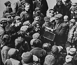 Fotografia czarno-biała. Zdjęcie wykonano z góry – z okna lub balkonu – ku dołowi, poziomowi ulicy – przedstawia to, co dzieje się poniżej – mężczyznę i zebraną wokół niego grupę osób, głównie dzieci i młodzieży. Mężczyzna – Jankiel Herszkowicz – stoi po prawej stronie kadru; jest ubrany w płaszcz, na głowie ma kaszkiet; trzyma znoszoną walizkę i kartkę, z której odczytuje, lub śpiewa, tekst. Wokół niego zgromadziły się dzieci i młodzież – publiczność słuchająca występu, w większości zapatrzeni w występującego – nieliczni spoglądają na osobę wykonującą zdjęcie. Zebrani są ubrani ciepło, większość ma nakrycia głów. Na ich odzieży wyraźnie widać naszyte gwiazdy Dawida.