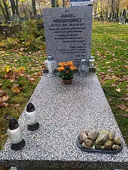 Fotografia kolorowa. Na zdjęciu część „B” Cmentarza Komunalego na Dołach w Łodzi zimą – widać pokrywający teren i groby śnieg. Na pierwszym planie, wykonany z tanich materiałów, nagrobek bez symboli religijnych. Pionowa część nagrobka z napisem „JAKUB HERSZKOWICZ UR. 22. VII. 1910 ZM. 25. III. 1972 CZEŚĆ JEGO PAMIĘCI”. Na nagrobku leży wiązanka sztucznych kwiatów i stoi znicz. Za nagrobkiem znajduje się pokryta śniegiem, szeroka aleja cmentarna a za nią kolejne nagrobki. Tło stanowią pozbawione liści drzewa.