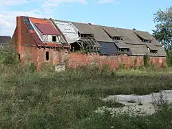 Na zdjęciu widać zrujnowany budynek gospodarczy z czerwonej cegły. Otwory okienne i drzwiowe w wielu miejscach są zamurowane. Dach drewniany, kryty najpewniej gontem, w wielu miejscach dziurawy. W niektórych miejscach dziury w dachu zostały załatane niepasującymi do reszty dachówkami, w innym miejscu zwykłą folią. Przed budynkiem rozciągają się gęste chaszcze.