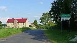 Zdjęcie przedstawia początek wsi Jastrzębnik. Z lewej strony widoczne są dwa domy. Z prawej strony znajduje się znak drogowy z nazwą wsi, informujący o początku terenu zabudowanego, oraz drzewa