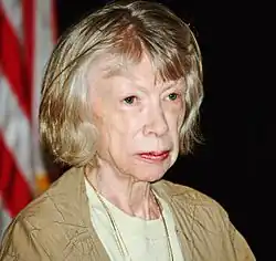 Zdjęcie portretowe Joan Didion. Starsza kobieta z włosami przyciętymi nad ramionami i prostą grzywką. W tle fragment flagi z białymi i czerwonymi pasami.