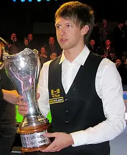 Zdjęcie portretowe od pasa. Judd po zawodach trzyma w rękach duży, metalowy puchar. Jest ubrany w białą koszulę i czarną kamizelkę. Włosy zaczesane do przodu. Za nim widać fragment zielnego sukna i publiczność.