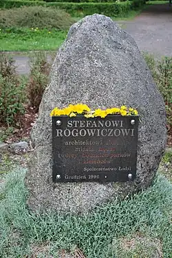 Zdjęcie kolorowe. Fotografia przedstawia szary, ustawiony pionowo, wśród parko wych alejek głaz narzutowy. Do kamienia jest przytwierdzona ciemna tablica pamiątkowa z napisem „STEFANOWI ROGOWICZOWI architektowi zieleni miasta Łodzi twórcy Łódzkich parków i zieleńców Społeczeństwo Łodzi Grudzień 1996”. Między górną krawędź tablicy, a głaz, do którego jest przytwierdzona, zatknięto na całej szerokości, kwitnące na żółto, kwiaty. W tle zdjęcia widać parkowe rośliny i alejki.