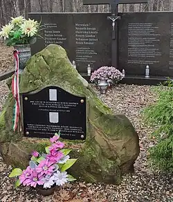 Zdjęcie zbiorowej mogiły w lesie Murckowskim. Ziemię pokrywają jesienne liście. Zdjęcie zrobione od boku po skosie. W pierwszym planie głaz o kształcie zbliżonym do ostrosłupa. Na nim czarna tabliczka z godłami Węgier i Polski oraz napisami w tych językach. Za nim znajduje się duża czarna kamienna płyta z wygrawerowanymi napisami. Gdzieniegdzie stoją wazony z kwiatami oraz znicze