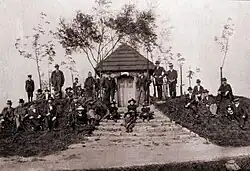 Czarno-białe archiwalne zdjęcie przedstawia nieduża kaplicę na wzniesieniu. Kaplica nakryta jest dachem pokrytym dachówką. Do kaplicy prowadzą szerokie schody. Przy kaplicy, na schodach i wzniesieniu są elegancko ubrani ludzie: mężczyźni, kobiety i dzieci. Część z nich stoi, część siedzi na schodach lub na trawie. Za kapliczką rośnie kilka niewielkich drzew.