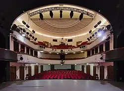 Zdjęcie wykonane ze sceny w kierunku na widownię. Scena otwiera się na widownię szerokim, ciemnym łukiem, od góry podwieszone są do niego reflektory. Na jasno oświetlonej widowni znajdują się rzędy czerwonych krzeseł. Ponad nimi są dwa poziomy balkonów. Do górnego balkonu, na całej jego długości, umocowane są reflektory.